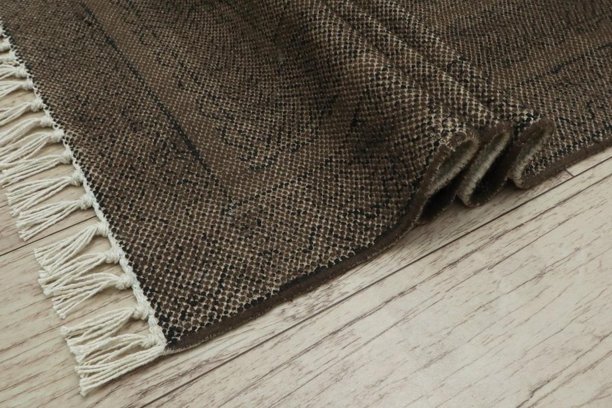 Corner of a brown fringed cotton rug on a wood floor